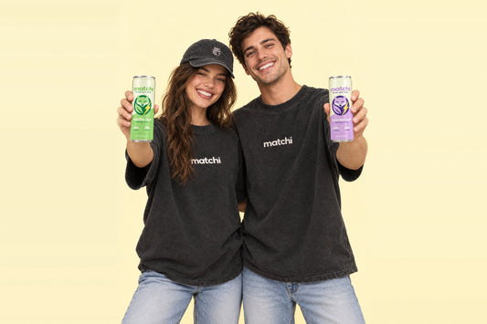 Two people in Matchi Den Issue 001 tees, one in a Matchi fox hat, holding up cans of Matchi Vanilla Oat and Lavender Oat Canned Matcha Latte with Protein